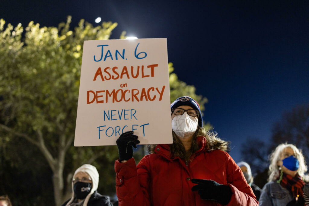 A woman holds a sign at a candlelight vigil that says "Jan. 6 Assault on Democracy, Never Forget". 