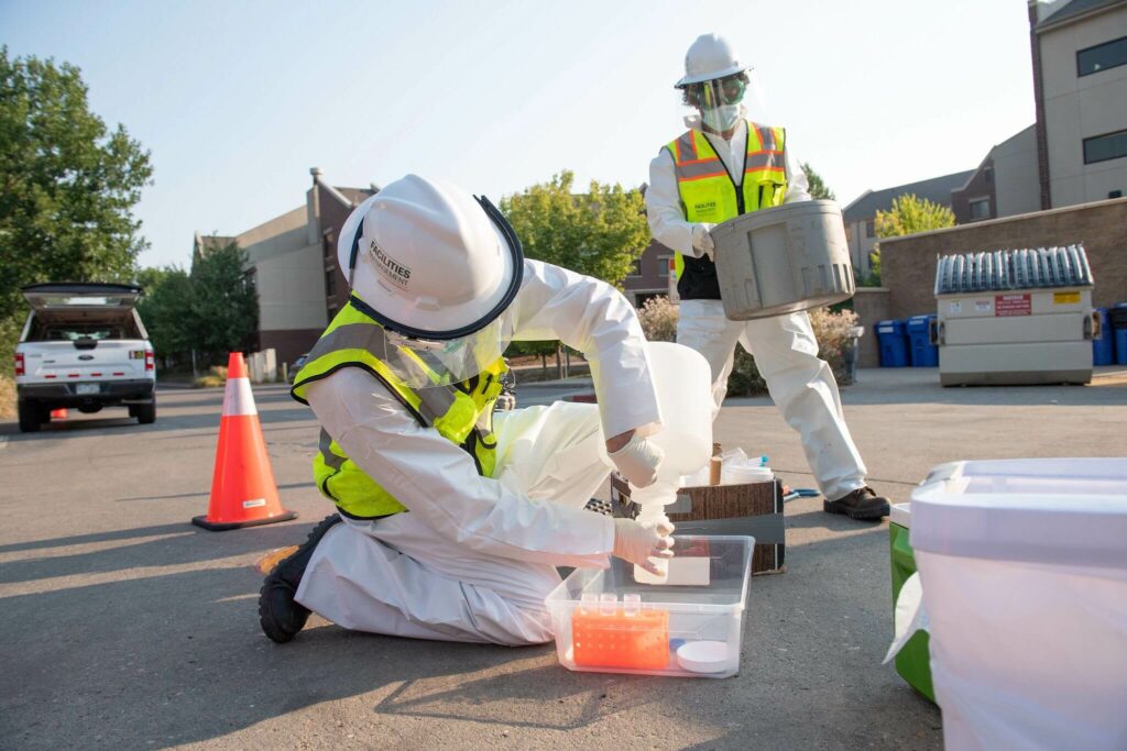 Two people outside in full protective gear place waste water into test tubes.