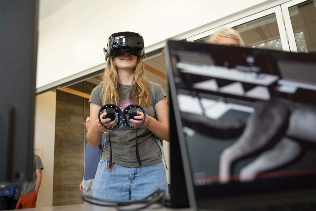 A woman wears a VR headset and holds controllers in both hands while in the foreground a monitor shows an animal laying on a table in the virtual reality space.