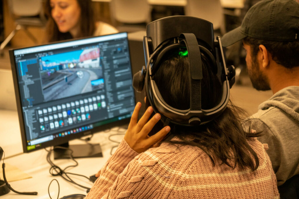 Two students sit at a computer with a virtual reality program on the screen. One wears a VR headset.