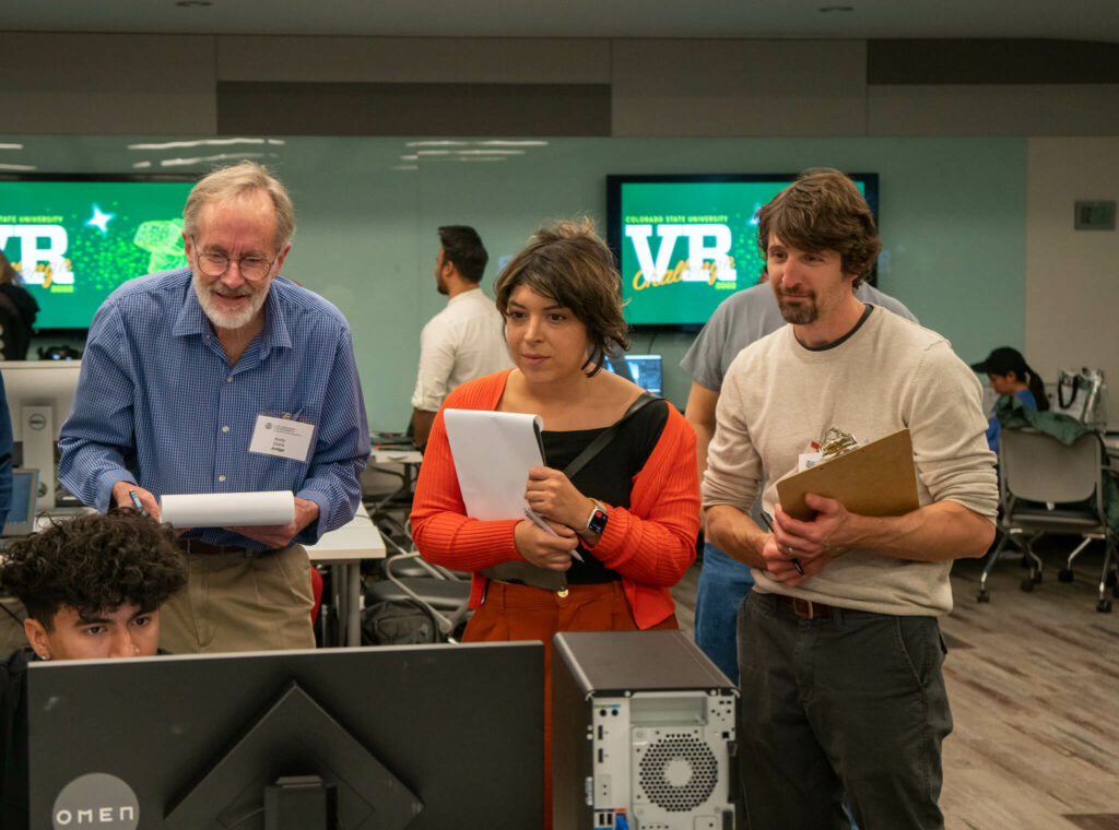 Three judges look at a student's computer screen while holding clipboards.