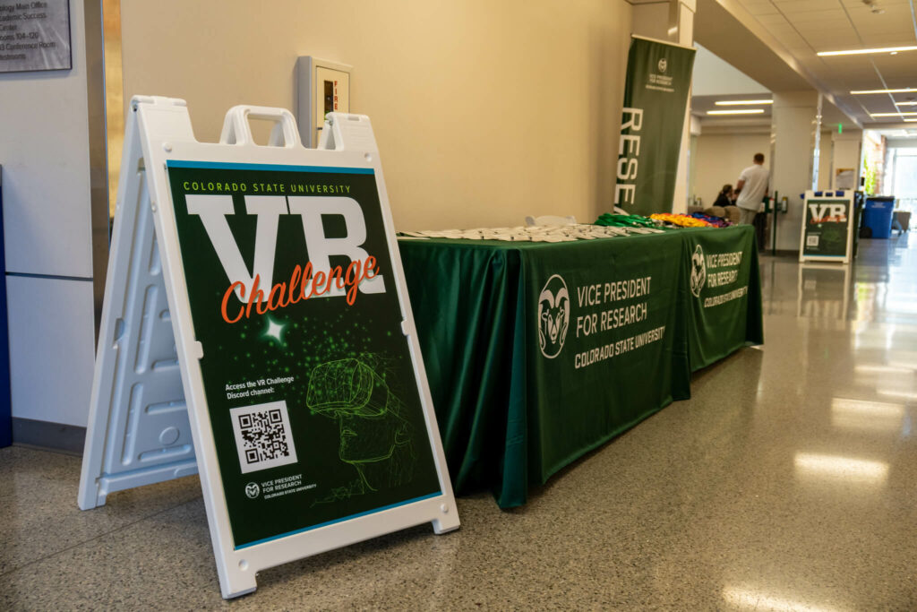 A sandwich board sign reading" Colorado State University VR Challenge" in front of a check-in table with nametags and swag.